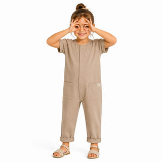 A young girl wearing a short-sleeve beige jumpsuit with front pockets, standing against a plain white background and making a binocular gesture with their hands.