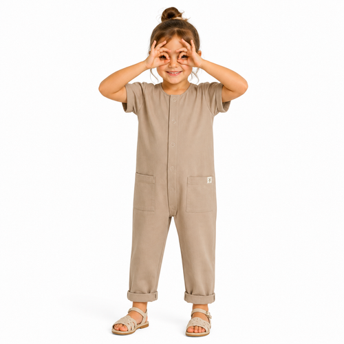A young girl wearing a short-sleeve beige jumpsuit with front pockets, standing against a plain white background and making a binocular gesture with their hands.