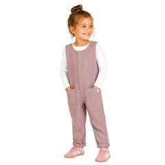A young girl with her hair in a bun stands against a white studio background wearing a dusty rose corduroy overall jumpsuit over a white long-sleeve shirt, paired with white socks and matching pink trainers.