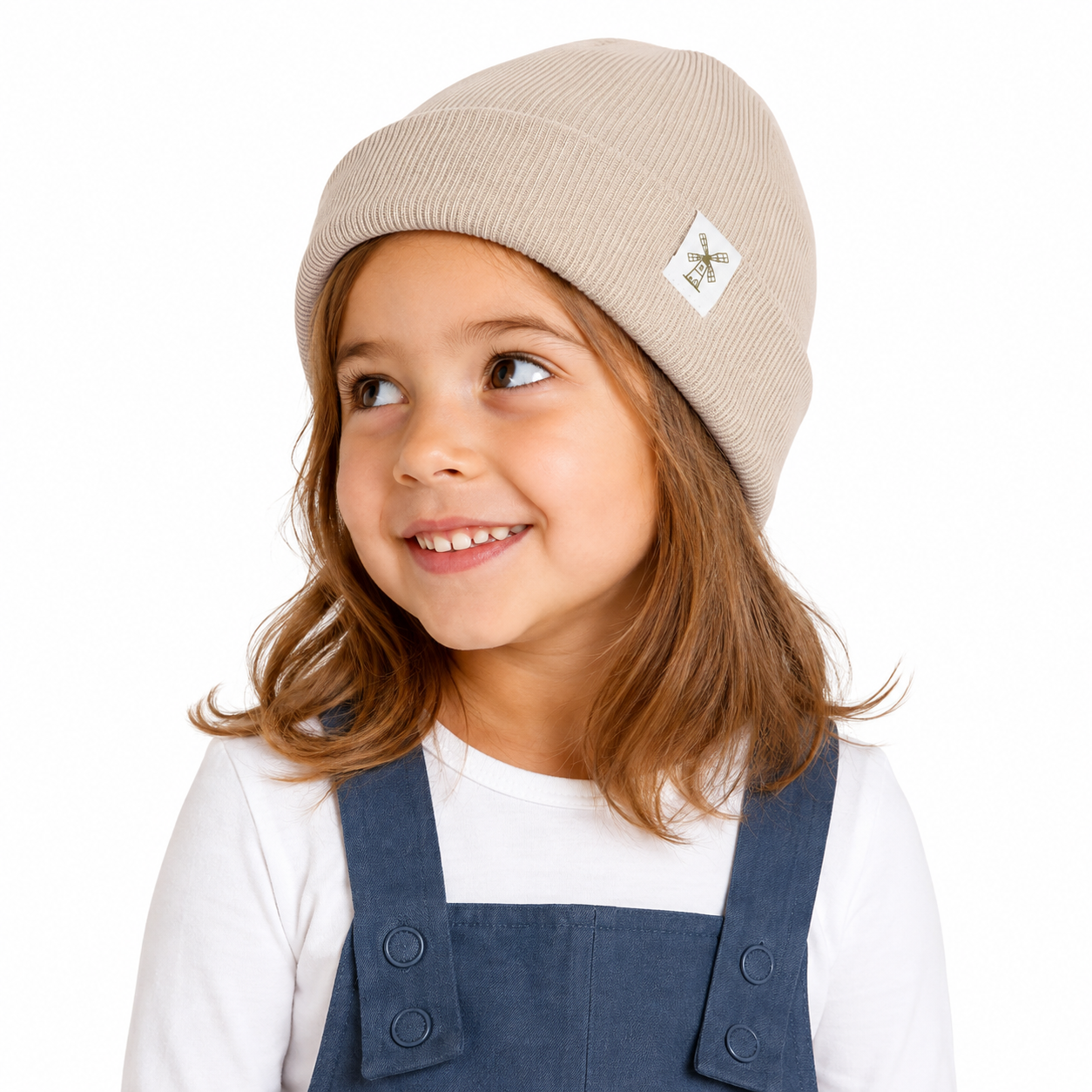 A young girl with brown hair wearing a beige ribbed knit beanie with a small white logo patch and navy blue denim overalls over a white long-sleeve shirt, photographed against a solid white background.