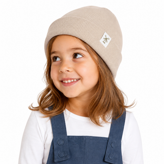 A young girl with brown hair wearing a beige ribbed knit beanie with a small white logo patch and navy blue denim overalls over a white long-sleeve shirt, photographed against a solid white background.