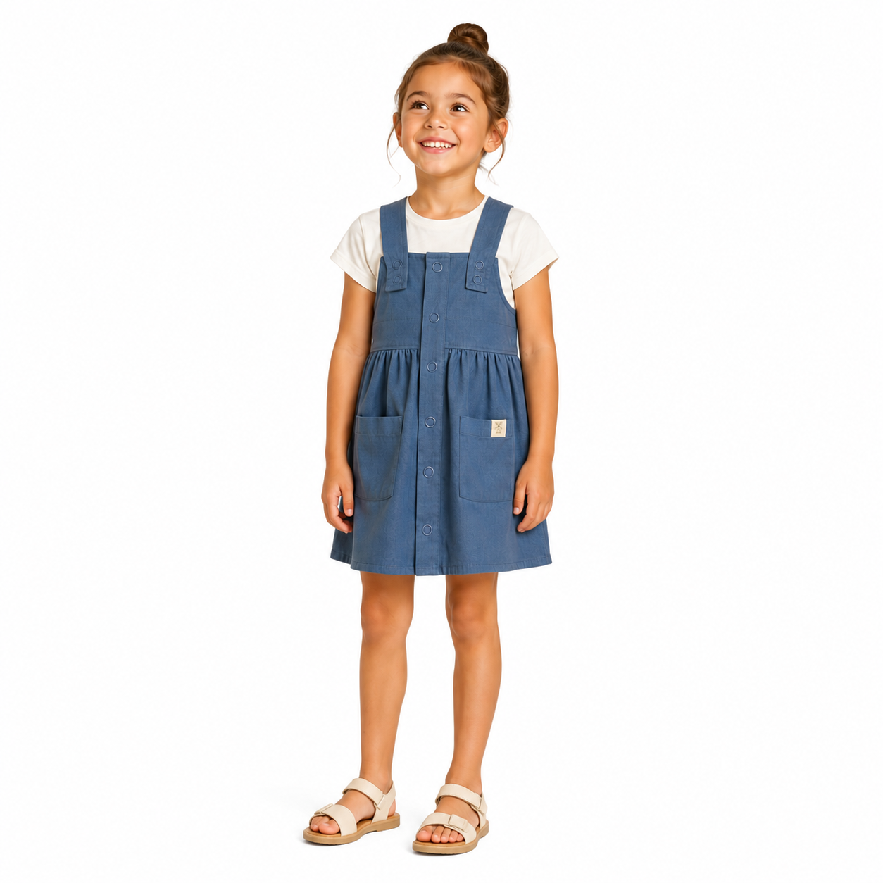 A young girl with her hair in a bun stands against a white background wearing a blue cotton dungaree dress over a white t-shirt and light-coloured sandals.