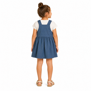 A young girl with her hair in a bun is shown from the back, wearing a blue dungaree-style pinafore dress over a white short-sleeve t-shirt and beige sandals, against a plain white background.