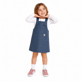 A young girl modeling a slate blue dungaree dress over a white long-sleeve shirt, paired with white socks and pink trainers, on a solid white background.