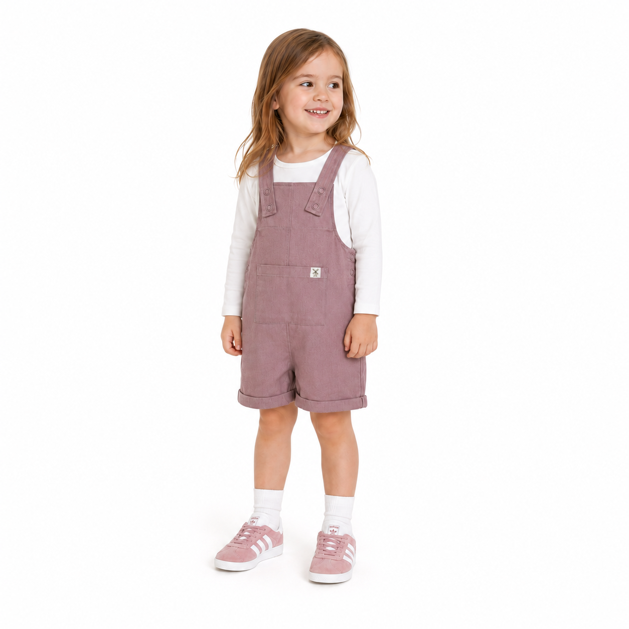 A young girl wearing dusty rose dungaree shorts over a white long-sleeve shirt, standing against a plain white background.