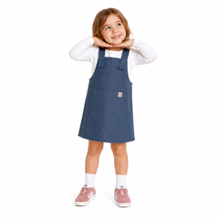 A young girl modeling a slate blue dungaree dress over a white long-sleeve shirt, paired with white socks and pink trainers, on a solid white background.