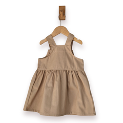 A sleeveless tan pinafore-style dress for a baby or toddler, hanging on a wooden hanger from a wall hook against a plain white background.