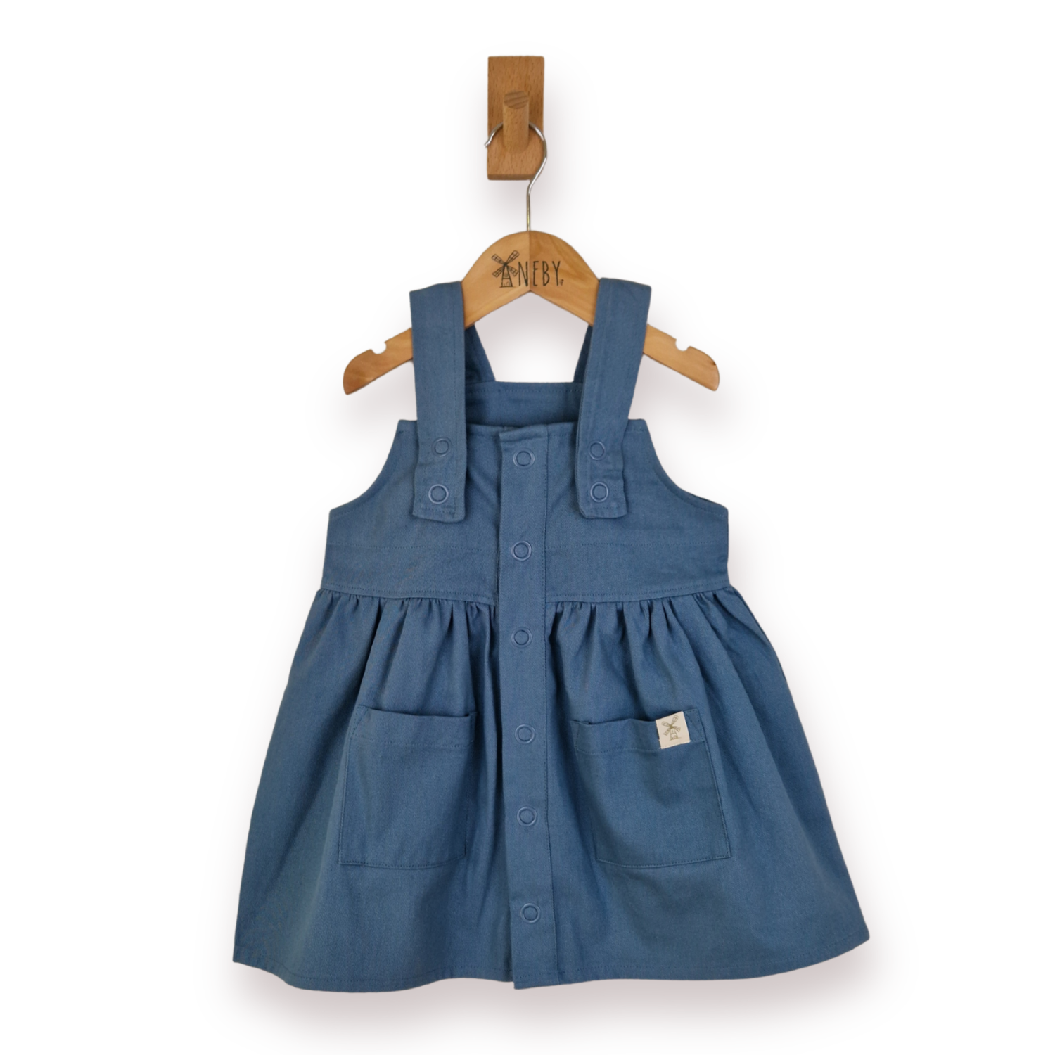 A blue twill pleated overall dress with front poppers and patch pockets, hanging on a wooden hanger against a white background.