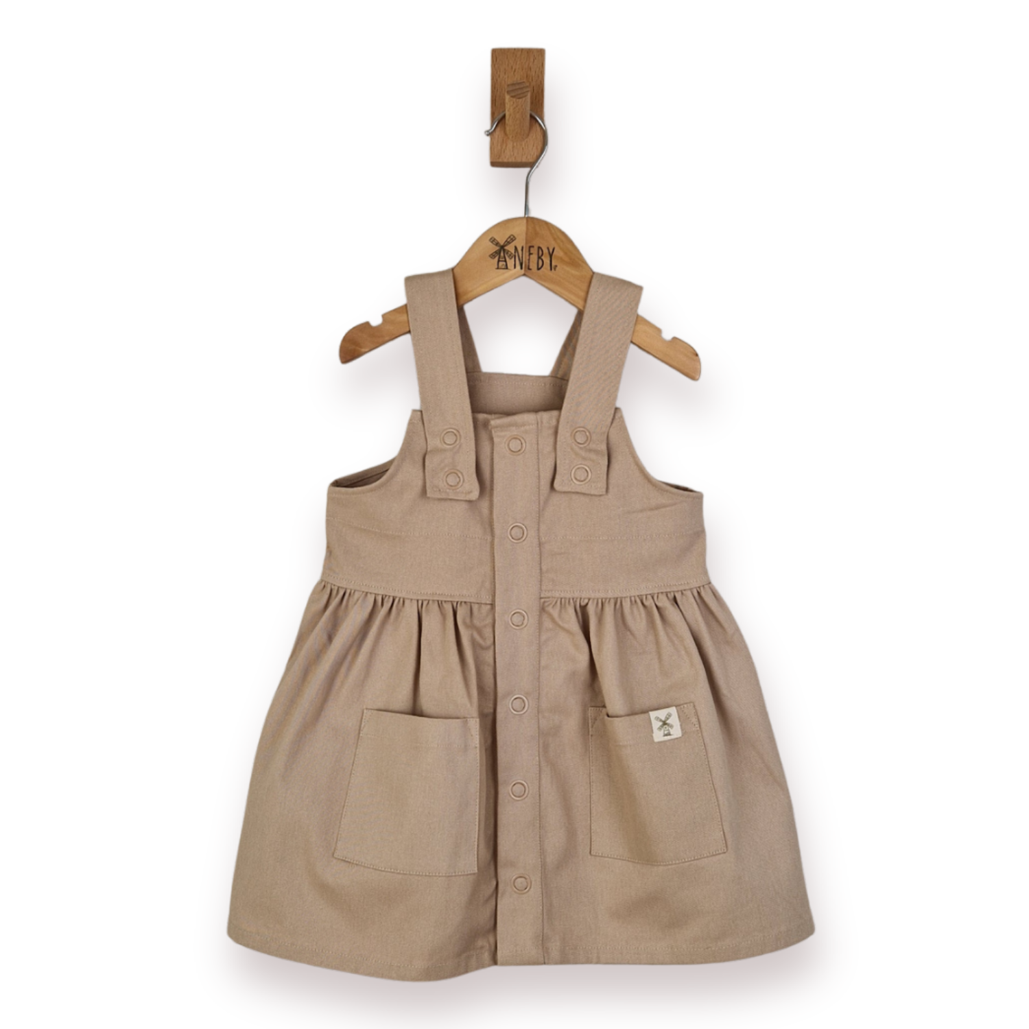 A toddler girl's beige cotton twill pinafore dress with front buttons and patch pockets, hanging on a wooden hanger from a wall peg against a white background.