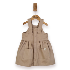 A toddler girl's beige cotton twill pinafore dress with front buttons and patch pockets, hanging on a wooden hanger from a wall peg against a white background.