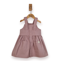 A dusty rose pink cotton twill pinafore dress for toddlers, featuring a button-down front, buttoned shoulder straps, and front patch pockets, hanging on a wooden hanger against a white background.