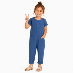 A young girl with her hair in a bun stands against a light gray background, wearing a short-sleeve blue jumpsuit with front pockets and button details, paired with beige sandals.