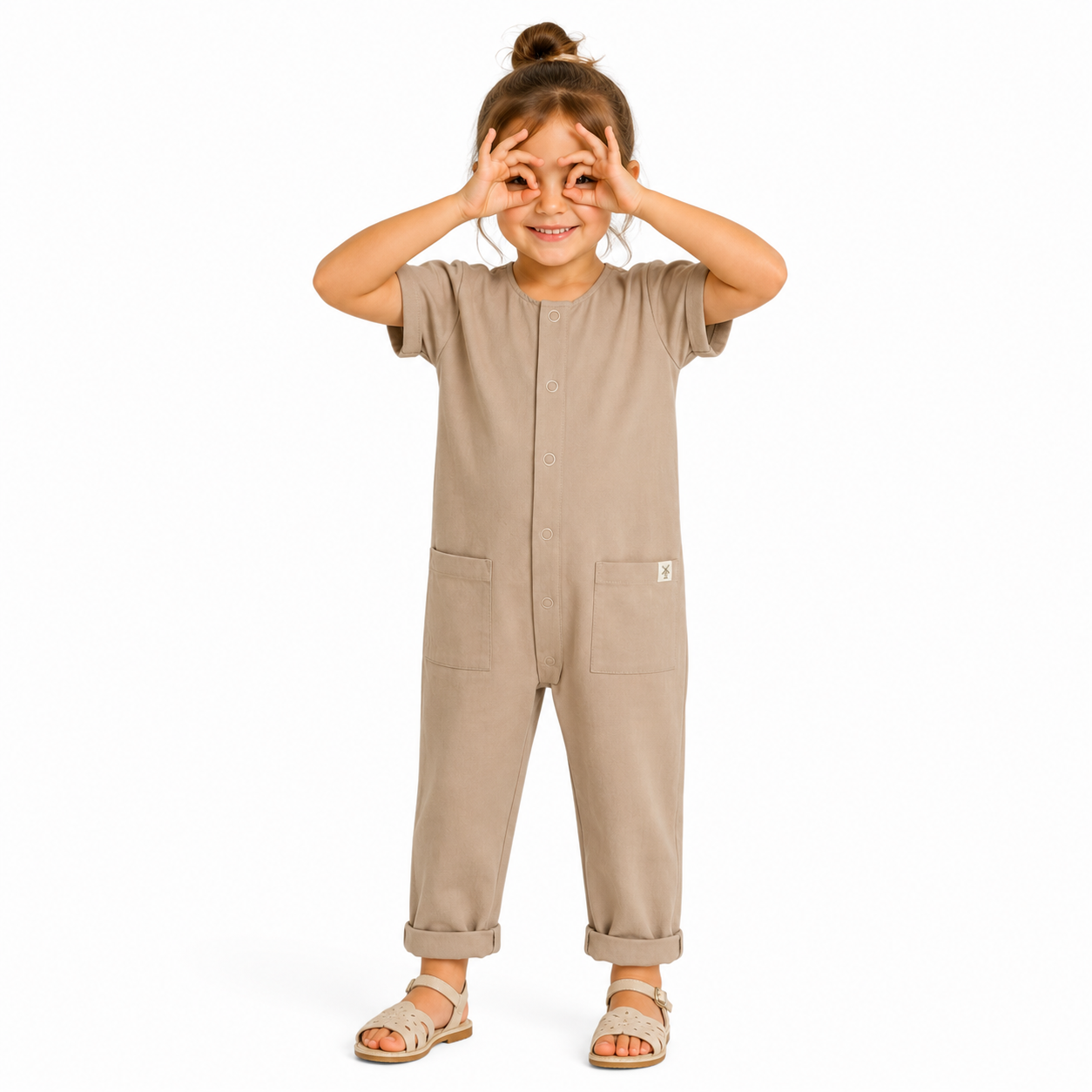 A young girl wearing a short-sleeve beige jumpsuit with front pockets, standing against a plain white background and making a binocular gesture with their hands.