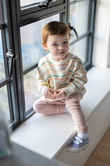 Model sitting against a window wearing striped sweatshirt and pink trousers