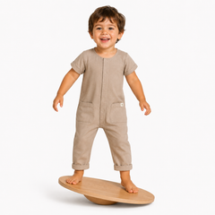 A smiling toddler stands barefoot on a wooden balance board, wearing a short-sleeve ecru jumpsuit with front pockets against a clean white background.