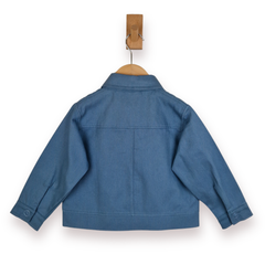 A blue twill cotton jacket shown from the back, hanging on a wooden hanger with a wooden peg hook above it, against a plain white background.