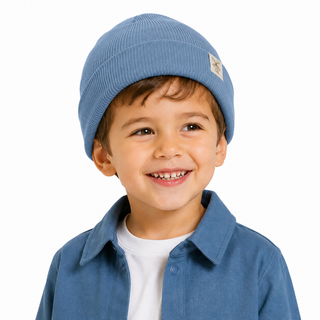 A smiling young boy wearing a light blue ribbed knit beanie with a small ecru brand patch and a matching blue button-up shacket over a white t-shirt, photographed against a clean white background.