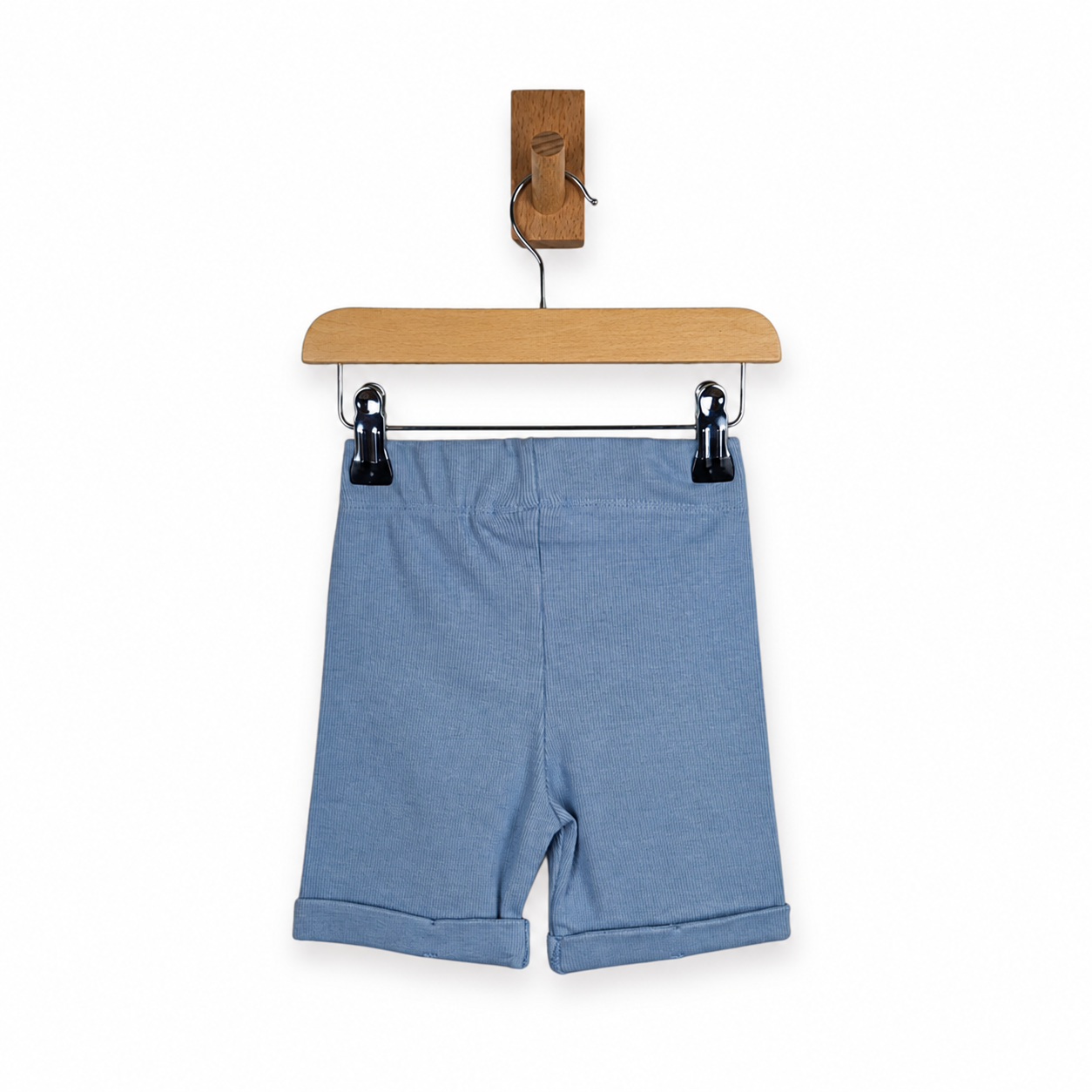 Dusty blue rib cotton knit shorts hanging from black clips on a wooden hanger with a wooden wall hook, against a plain white background.