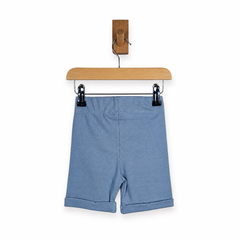 Dusty blue rib cotton knit shorts hanging from black clips on a wooden hanger with a wooden wall hook, against a plain white background.