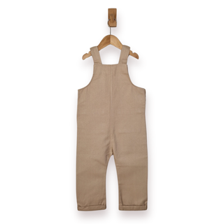 A pair of toddler beige dungaree overalls in a light tan colour, hanging on a wooden hanger from a wall hook against a white background.