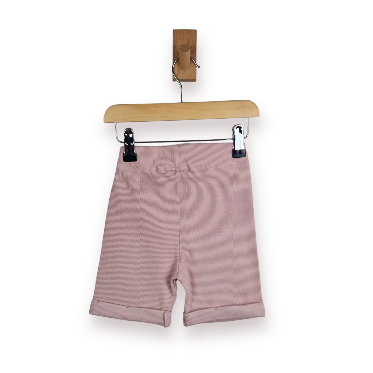A pair of dusty pink ribbed knit shorts with rolled cuffs, hanging from a wooden hanger with black clips against a plain white background.