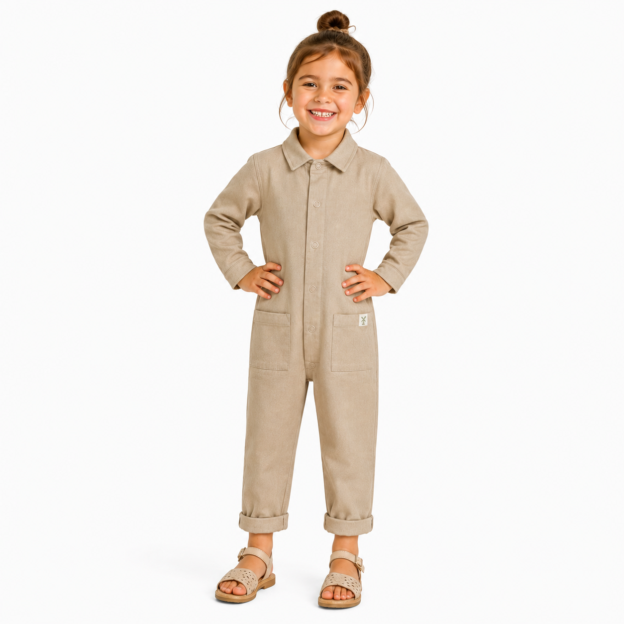 A young girl with her hair in a bun stands wearing a light beige long-sleeve Boilersuit with a collar, button front, and front pockets, paired with brown sandals, against a plain white background.