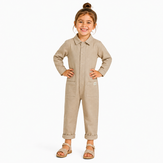 A young girl with her hair in a bun stands wearing a light beige long-sleeve Boilersuit with a collar, button front, and front pockets, paired with brown sandals, against a plain white background.