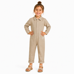 A young girl with her hair in a bun stands wearing a light beige long-sleeve Boilersuit with a collar, button front, and front pockets, paired with brown sandals, against a plain white background.