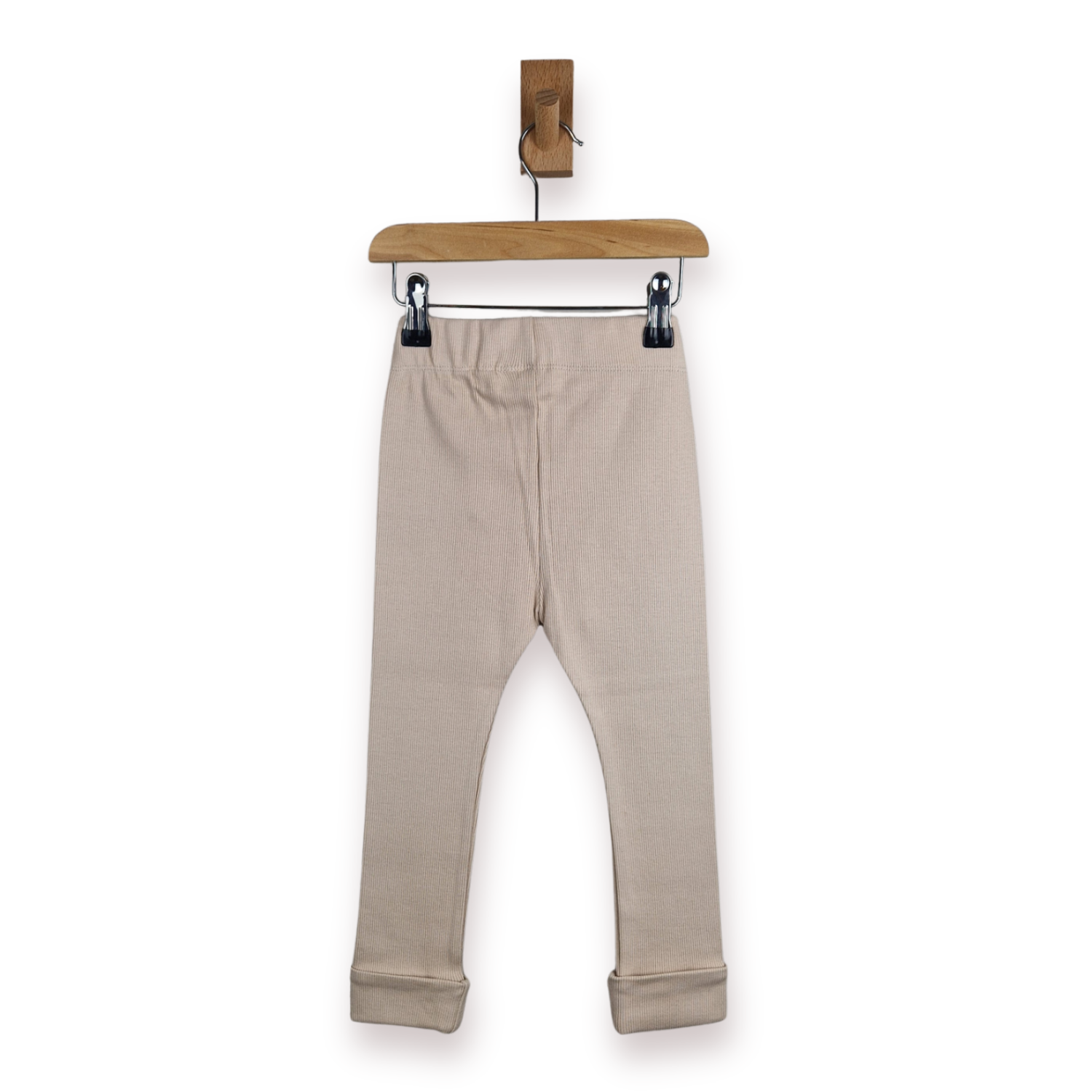 A pair of kids light beige leggings with rolled cuffs, hanging from a wooden hanger with black clips against a plain white background.