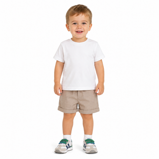 A toddler standing against a plain white background, wearing a plain white short-sleeve t-shirt, beige shorts, white socks, and grey trainers with green accents.