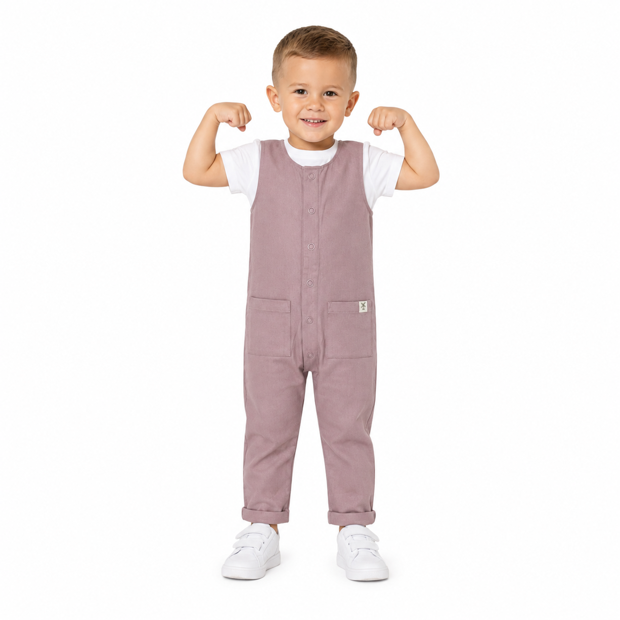 A young boy modelling a pink overall jumpsuit over a white t-shirt and white trainers, posing with his arms flexed against a solid white background.
