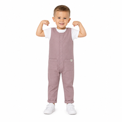 A young boy modelling a pink overall jumpsuit over a white t-shirt and white trainers, posing with his arms flexed against a solid white background.