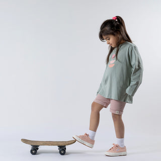 Girl in green long sleeved t-shirt with abstract print on the front, wearing pink shorts standing on a skateboard