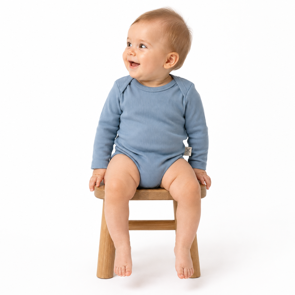 A baby wearing a dusty blue long-sleeve bodysuit sits on a small wooden stool against a clean white background.