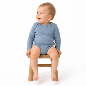 A baby wearing a dusty blue long-sleeve bodysuit sits on a small wooden stool against a clean white background.
