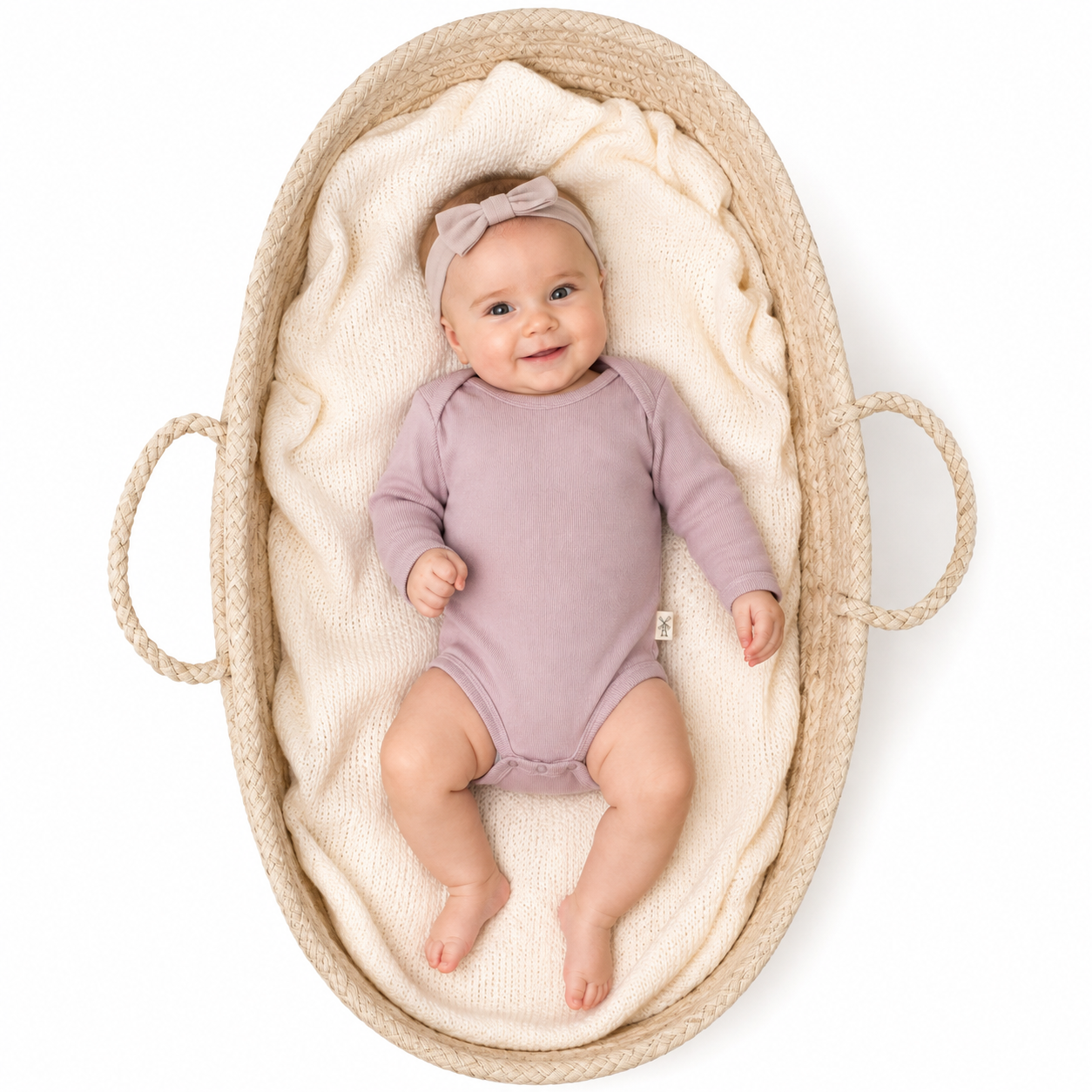 A baby wearing a pink long-sleeve bodysuit and a matching bow headband, lying on a cream knit blanket inside a woven basket with rope handles, photographed from above on a white background.