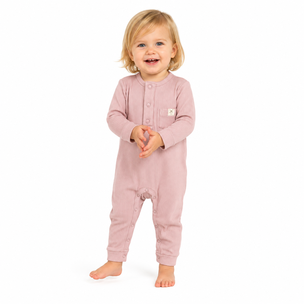A toddler standing barefoot against a plain white background, wearing a dusty pink long-sleeve footless one-piece jumpsuit with a buttoned chest and a small white square logo patch.