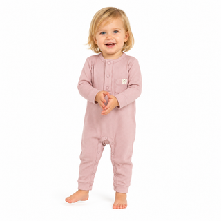 A toddler standing barefoot against a plain white background, wearing a dusty pink long-sleeve footless one-piece jumpsuit with a buttoned chest and a small white square logo patch.
