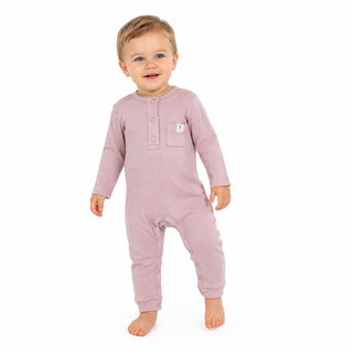 A toddler standing barefoot against a plain white background, wearing a dusty pink long-sleeve footless one-piece romper with a three-button placket and a small chest pocket.
