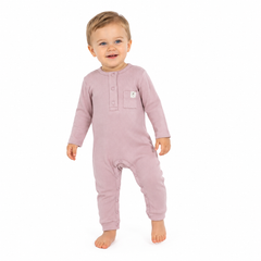 A toddler standing barefoot against a plain white background, wearing a dusty pink long-sleeve footless one-piece romper with a three-button placket and a small chest pocket.