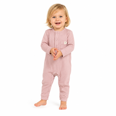 A toddler standing barefoot against a plain white background, wearing a dusty pink long-sleeve footless one-piece jumpsuit with a buttoned chest and a small white square logo patch.