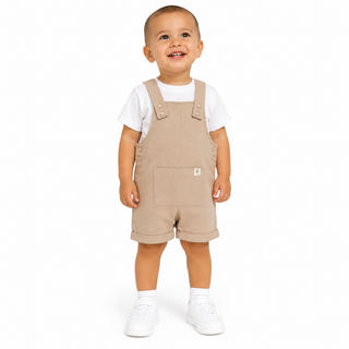 A toddler standing against a white studio background wearing beige corduroy overall shorts over a white t-shirt and white sneakers.