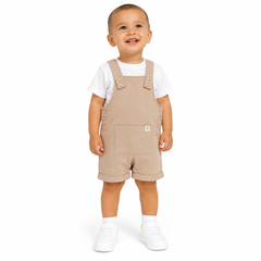 A toddler standing against a white studio background wearing beige corduroy overall shorts over a white t-shirt and white sneakers.