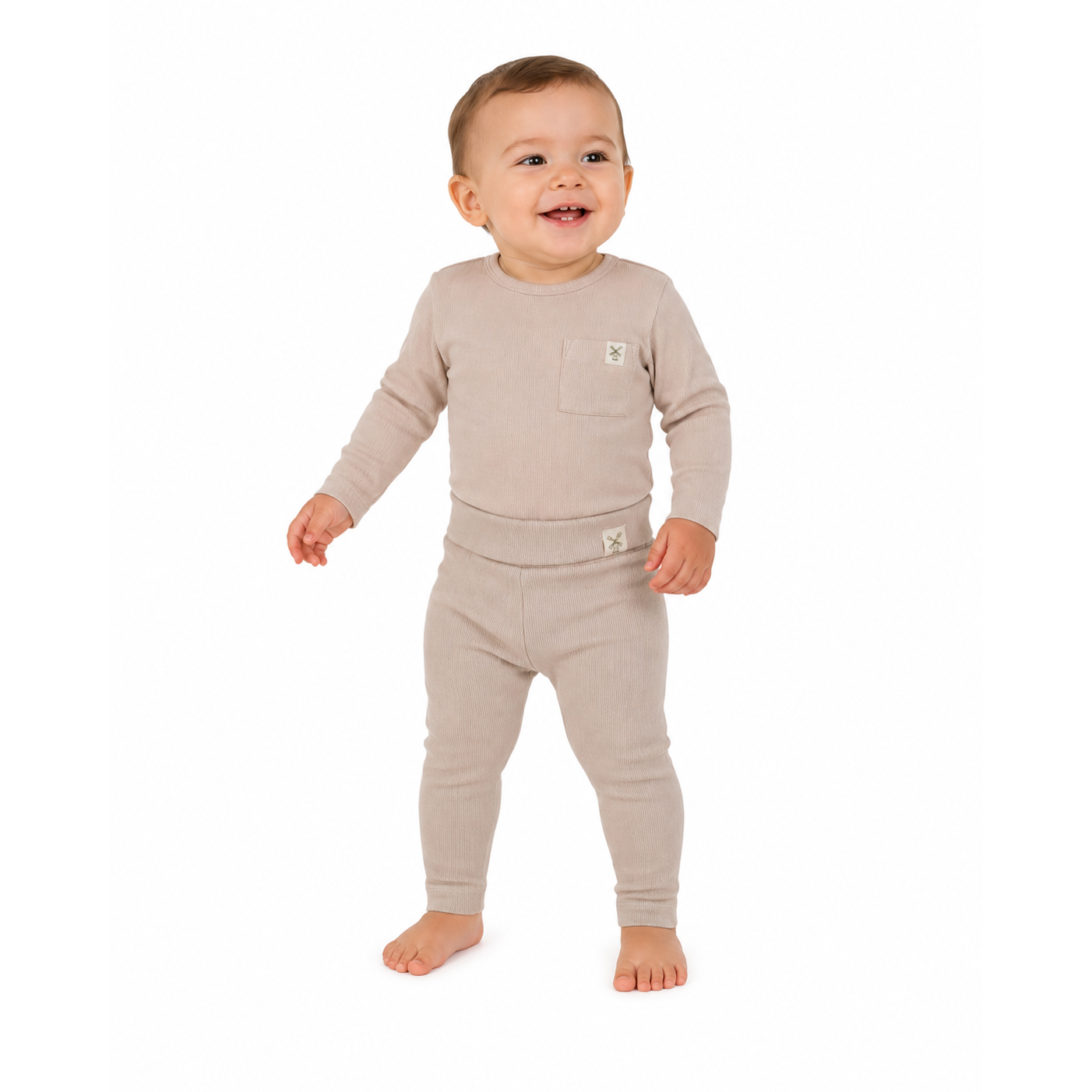 A smiling baby standing barefoot in a light beige knit two-piece outfit with a long-sleeve top and matching leggings, photographed against a plain white background.