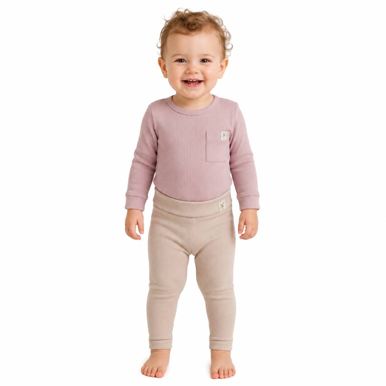 A smiling toddler standing barefoot against a plain white background, wearing a ribbed long-sleeve top in dusty mauve with a chest pocket and matching beige leggings.
