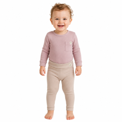 A smiling toddler standing barefoot against a plain white background, wearing a ribbed long-sleeve top in dusty mauve with a chest pocket and matching beige leggings.