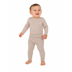 A smiling baby standing barefoot in a light beige knit two-piece outfit with a long-sleeve top and matching leggings, photographed against a plain white background.