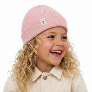 A young child with long curly blonde hair wearing a light pink ribbed knit beanie with a small white embroidered logo patch, paired with a cream-colored collared shirt, set against a solid white background.