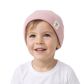 A smiling young child wearing a light pink ribbed knit beanie with a small white brand patch on the side, photographed against a clean white background.