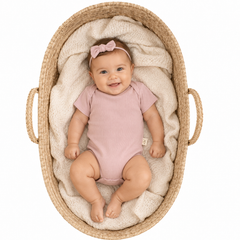 A baby wearing a dusty pink long-sleeve bodysuit and a matching bow headband, lying on a cream knit blanket inside a woven Moses basket with rope handles, photographed from above on a white background.